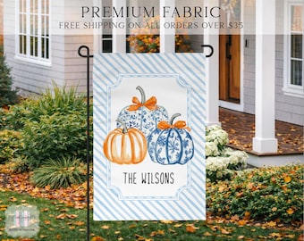 May include: A rectangular garden flag featuring a light blue and white diagonal striped background. The center displays three pumpkins, one solid orange and two with blue and white floral patterns, each topped with an orange bow. The text THE WILSONS is printed below.