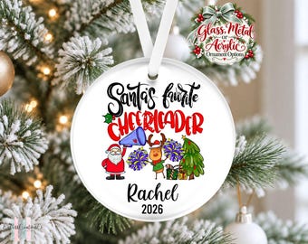 May include: A round white Christmas ornament featuring the text Santa's favorite CHEERLEADER, the name Rachel, and the year 2026. The design includes a cartoon Santa, a reindeer holding purple and yellow pom-poms, a megaphone, and a tree.