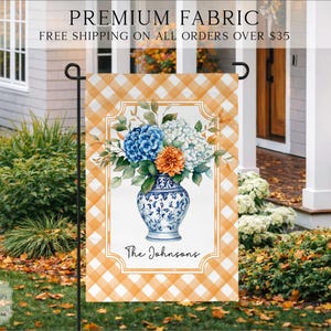 May include: A rectangular garden flag featuring an orange and white buffalo check pattern. The center displays a blue and white porcelain vase filled with blue, white, and orange flowers. The text The Johnsons is printed in black script.