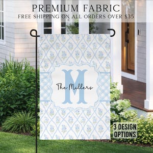 May include: A light blue and white garden flag with a floral pattern and the text "The Millers". The flag has a light blue border and a large letter "M". The flag is displayed outdoors.