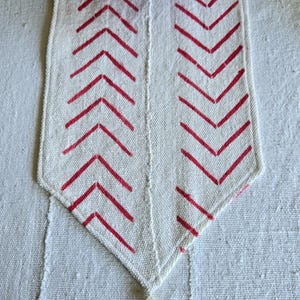Könnte beinhalten: Cremefarbener Tischläufer mit rotem Zickzackmuster und Quaste. Der Läufer besteht aus gewebtem Stoff und hat ein spitzes Ende. Ein roter Teller mit gewelltem Rand und ein Blumengewebe sind oben sichtbar.