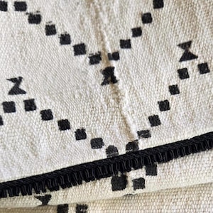 May include: Close-up of a cream-colored textile with a geometric pattern of black squares and arrow-like shapes. The edge is trimmed with a black fringe. The fabric appears to be woven.