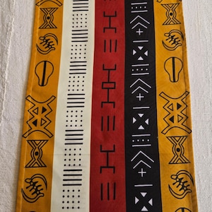 Puede incluir: Camino de mesa rectangular con un patrón repetido de rayas verticales en amarillo mostaza, blanco, rojo y negro. El diseño incluye formas y símbolos geométricos. El camino de mesa es probablemente de tela.