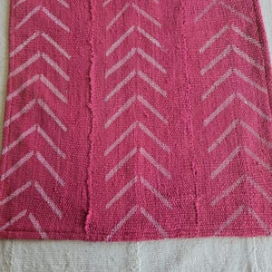 May include: A rectangular, woven rug in a vibrant fuchsia color. The rug features a repeating pattern of white chevron arrows. The texture of the rug is visible, suggesting a handmade or natural fiber construction. The rug is placed on a white surface.