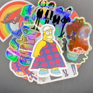 May include: A collection of colorful stickers featuring a cartoon bear, a cartoon character wearing a dress, a rainbow, a car, and several hand gestures. The stickers are holographic and have a shiny finish.