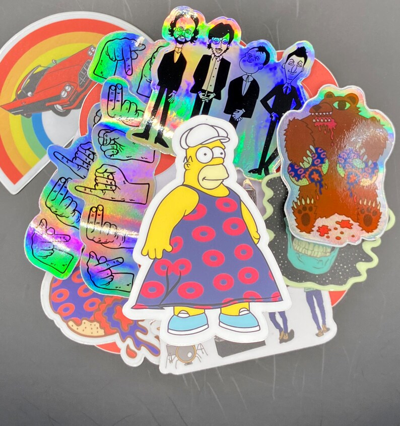 May include: A collection of colorful stickers featuring a cartoon bear, a cartoon character wearing a dress, a rainbow, a car, and several hand gestures. The stickers are holographic and have a shiny finish.