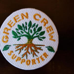 May include: A circular white embroidered patch featuring a green tree with visible roots in the center. The text "GREEN CREW" is arched across the top and "SUPPORTER" is arched across the bottom in yellow thread. Two small green leaves are placed on the left and right sides.