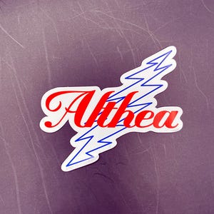May include: A die-cut sticker featuring the word Althea written in a bold red script font. Behind the text is a blue outlined lightning bolt shape. The sticker has a white border and is set against a dark purple background with subtle texture.