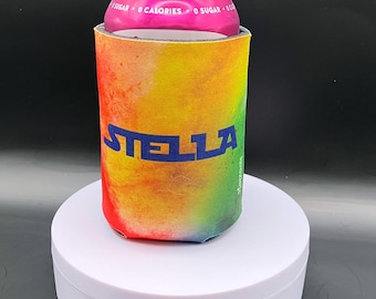 May include: A rainbow tie-dye can cooler with the word "STELLA" in blue lettering. The cooler is made of neoprene and is designed to keep drinks cold.