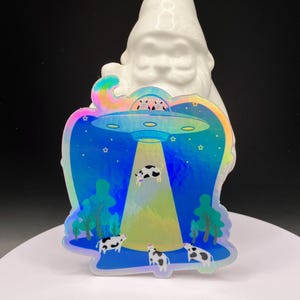 May include: A holographic sticker featuring a blue night sky with stars and a flying saucer beaming down a light. A cow is being abducted by the UFO, while other cows stand on the ground near trees. The sticker has a reflective, iridescent finish.