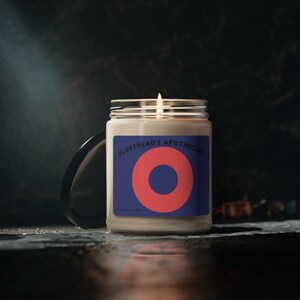 Op de afbeelding: Een beige kaars met een blauw en rood etiket met de tekst "Fluffhead's Apothecary". Het etiket heeft een grote rode cirkel met een kleinere blauwe cirkel erin.