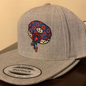 Puede incluir: Gorra snapback gris con visera plana. Presenta un diseño bordado de una rosquilla azul y roja con un mordisco. La etiqueta dice "The Authentic Snapback Yupoong Since 1974".