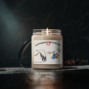 Peut inclure: Une bougie en soja beige avec une étiquette blanche illustrant un dessin animé d'une cigogne portant un bébé dans une couverture bleue. L'étiquette porte les inscriptions "Fluffhead's Apothecary" et "Check it out!" en texte noir.