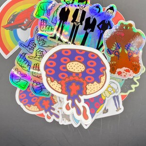 May include: A collection of colorful stickers featuring a donut with red and purple frosting, a bear wearing a donut-patterned shirt, a rainbow with a red car, and a group of men in suits. The stickers are holographic and have a glossy finish.