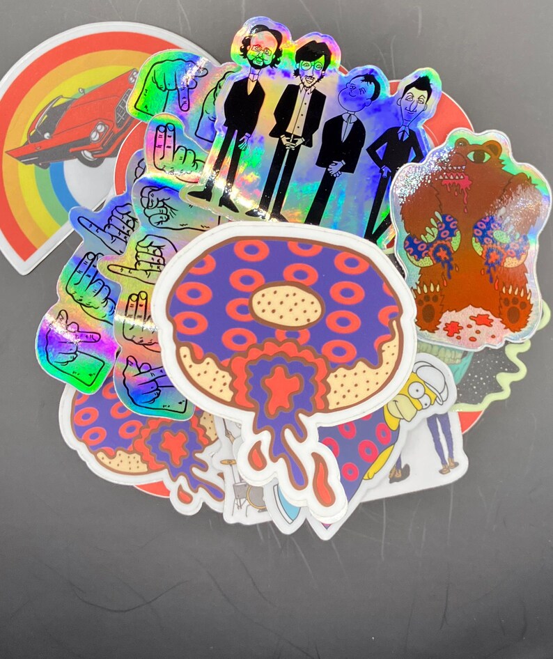 May include: A collection of colorful stickers featuring a donut with red and purple frosting, a bear wearing a donut-patterned shirt, a rainbow with a red car, and a group of men in suits. The stickers are holographic and have a glossy finish.