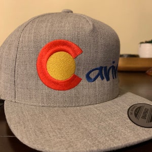 May include: A heather gray snapback cap featuring an embroidered red and yellow circular logo on the front panel. The word "ariv" is embroidered in blue script next to the logo. A circular black sticker with white text is attached to the flat brim.