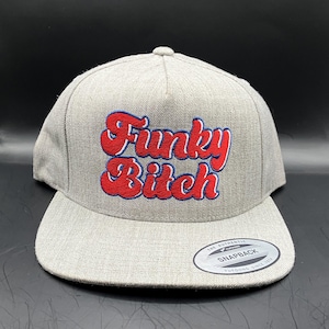 May include: A heather gray snapback cap featuring the embroidered text Funky Bitch in red with a blue outline on the front panel. The hat has a flat brim and a circular sticker on the right side that reads The Authentic Yupoong Snapback.