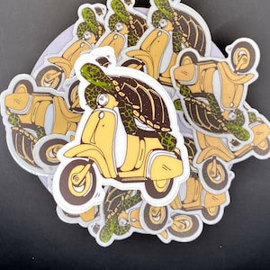 May include: A stack of yellow and white scooter stickers with a green turtle riding on the scooter. The stickers are on a gray background.