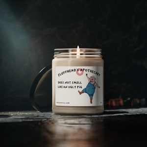 Op de afbeelding: Een bruine sojawas-kaars in een glazen pot met een roze en wit etiket waarop "Fluffhead's Apothecary" en "Ruikt niet naar een lelijk varken" staat. Het etiket toont ook een cartoon-varken dat een overall draagt.