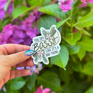 Può includere: Un adesivo bianco con il testo "YOU SAY BOSS LIKE IT'S A BAD THING" in nero, con accenti floreali e di foglie. L'adesivo è tenuto su uno sfondo di foglie verdi e fiori rosa.
