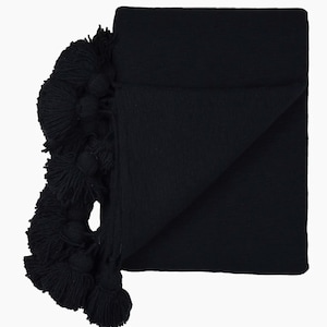 May include: A folded black throw blanket with a textured knit design. The blanket features a row of large, fluffy tassels along one edge. The throw is made of a soft material, perfect for adding warmth and style to any living space.