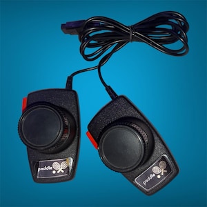 May include: A pair of black video game paddle controllers with a textured finish, each featuring a large circular dial and a red side button. A black cable connects both controllers to a single connector plug. Each unit displays a label with the word paddle.