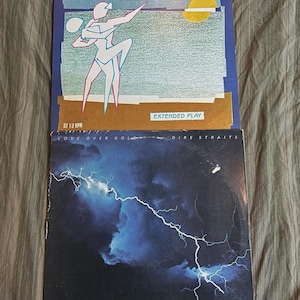 May include: Two Dire Straits vinyl record covers. The top cover is the Extended Play EP with a stylized dancing figure on a beach. The bottom cover is Love Over Gold, featuring a bright white lightning bolt across a dark blue cloudy sky.