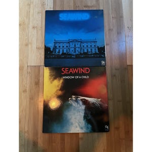 May include: Two Seawind vinyl record albums. The top cover features a blue-toned mansion with the text SEAWIND. The bottom cover, titled SEAWIND WINDOW OF A CHILD, displays a dark, fiery abstract image with a fish-like creature in the foreground.