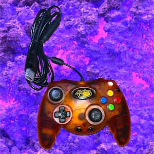 May include: A translucent amber-colored wired game controller featuring the Mad Catz logo in the center. The device includes a black directional pad, two black analog sticks, and four colored action buttons in blue, yellow, green, and red.