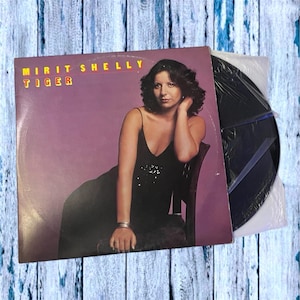 Pode incluir: Capa de álbum de vinil vintage para "Mirit Shelly Tiger". A capa apresenta uma mulher em um vestido preto, sentada em uma cadeira, com o título do álbum e o nome do artista em texto amarelo. O disco está em uma capa de plástico.