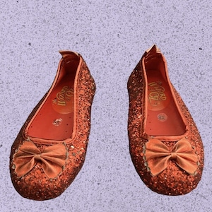 Op de afbeelding: Een paar rode glitterschoenen met een gouden strik en de tekst "The Wizard of Oz" aan de binnenkant.
