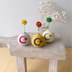 May include: Three felt ball vases with colorful silk flowers. The vases are white, yellow, and cream, each with a crescent moon and star design. The flowers are yellow, orange, and green. The vases are on a light wood table.