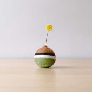 May include: A small, round, green, brown, black and white felt vase with a single yellow felt flower.