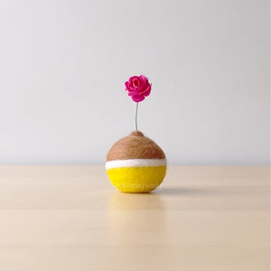 May include: A small, round, yellow and brown felt vase with a white stripe around the middle. A single pink felt flower sits on top of the vase.