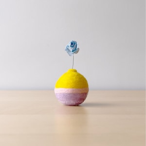May include: A small, yellow and pink felt ball vase with a single blue paper flower on top.