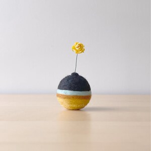 Puede incluir: Un pequeño jarrón redondo de fieltro negro, blanco y amarillo con una flor de papel amarilla en la parte superior.