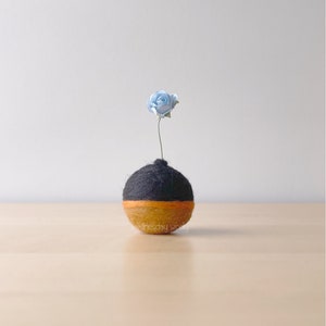 May include: A small, round, felted vase with a black top and orange bottom holds a single light blue flower.