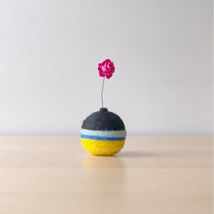 Könnte beinhalten: Eine kleine, runde, gelbe, blaue und schwarze Filzvase mit einer einzelnen rosa Blume.
