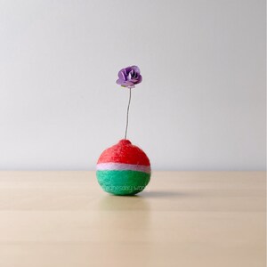 May include: A small, round, red, green, and white felt ball with a purple flower on top. The ball is sitting on a light wood surface.