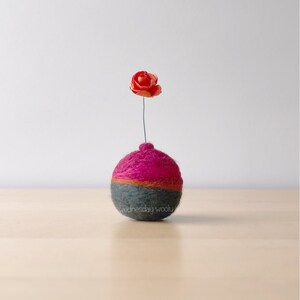Könnte beinhalten: Eine kleine, runde, gefilzte Vase in einem rosa und grauen Farbschema. Eine einzelne orangefarbene Blume ist in die Vase gestellt.