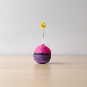 Könnte beinhalten: Eine kleine, runde Filzvase mit rosa Oberteil und lila Unterteil. Eine einzelne gelbe Filzblume sitzt oben auf der Vase.