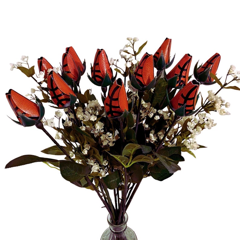 May include: A bouquet of orange and black basketball-themed roses in a clear glass vase. The roses are arranged in a vase with green leaves and white flowers.