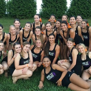 May include: A group of young women in black athletic outfits pose on a grassy field. The outfits include tank tops with the text "BOBCAT NATION" and matching shorts. Many have orange hair accessories. The image is taken outdoors.