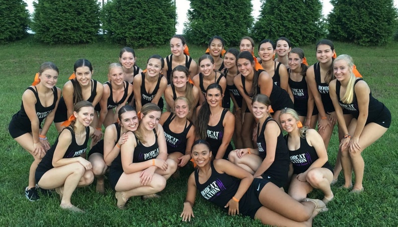 May include: A group of young women in black athletic outfits pose on a grassy field. The outfits include tank tops with the text "BOBCAT NATION" and matching shorts. Many have orange hair accessories. The image is taken outdoors.
