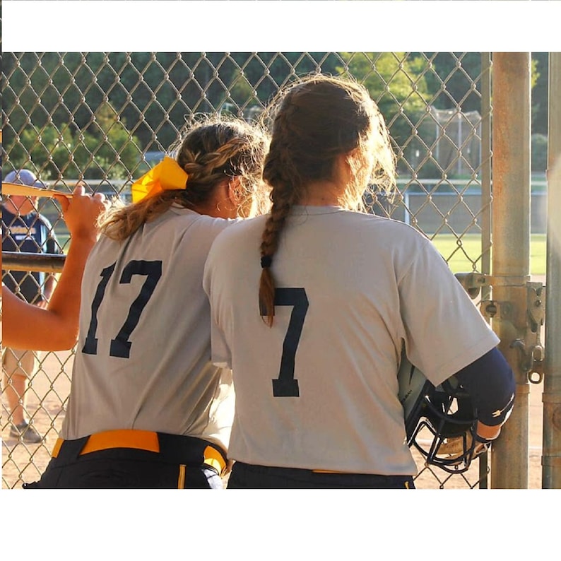 May include: Two softball players wearing light gray jerseys with navy blue numbers 17 and 7. One player has a yellow bow in her hair. The other player is holding a black helmet. They are standing near a chain-link fence.