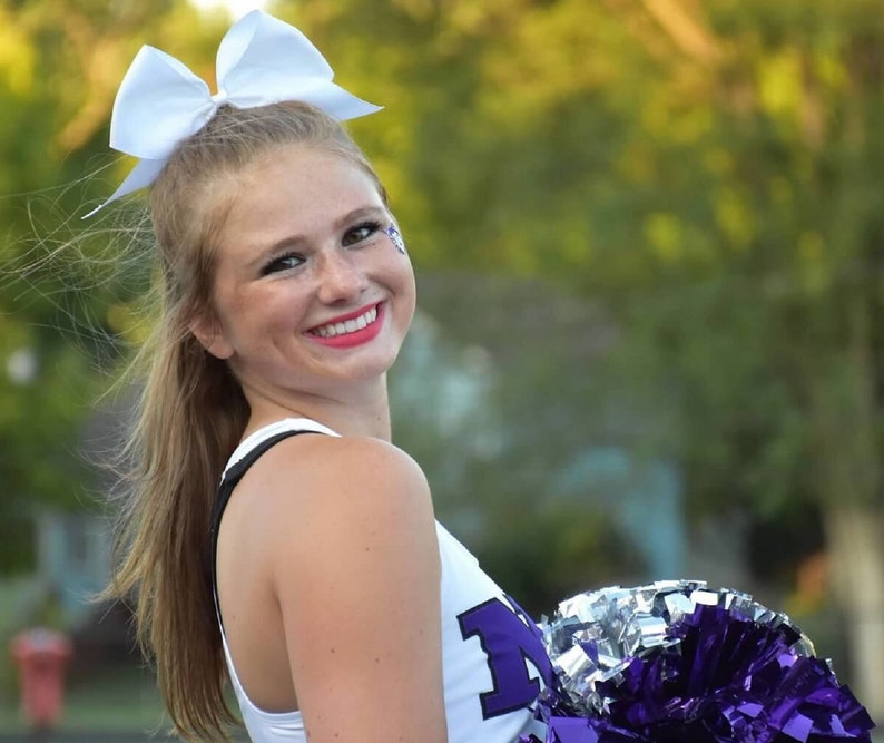 May include: A young woman with blonde hair wearing a white cheerleading uniform with purple lettering. She is smiling and holding a purple and silver pom-pom.