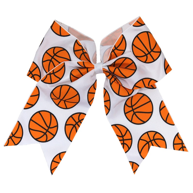 May include: A large hair bow made of white grosgrain ribbon featuring a repeating pattern of orange basketballs with black outlines. The bow is constructed with multiple loops and two long, pointed tails extending downward from the centre knot.
