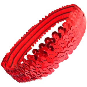 May include: A red sequin headband. The headband is made of small, shiny, circular sequins. The sequins are attached to a red elastic band. The headband is designed to be worn around the head. The sequins are closely packed together, creating a shimmering effect.