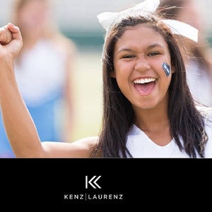 May include: A smiling person with dark hair and a white bow in their hair, wearing a white top, raises a fist in the air. A small face tattoo is visible. The logo "KENZ | LAURENZ" is at the bottom.