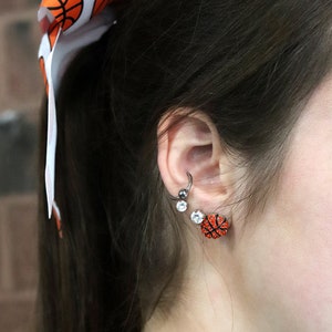 May include: A person with long dark hair wearing a basketball-themed hair bow and earrings. The earrings are small and feature a basketball design with rhinestones.